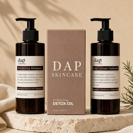 DAP Skincare pre-wash scalp detox oil set featuring two dark amber pump bottles with black caps and a central brown box, styled on a stone slab with a beige background.