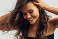 A close-up lifestyle shot of a smiling woman with long, wavy brown hair, wearing a black strapless top against a light gray background.