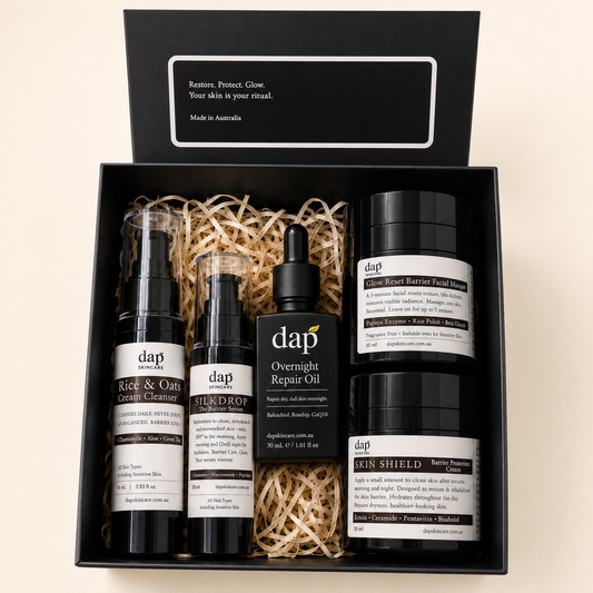 A Dap skincare gift set in a black presentation box with shredded paper filler, containing four black bottles and jars with white labels: RAE & OIL Cleanser, SILKDROP Repair Cream, Overnight Repair Oil, and SKIN SHIELD Barrier Repair Balm.